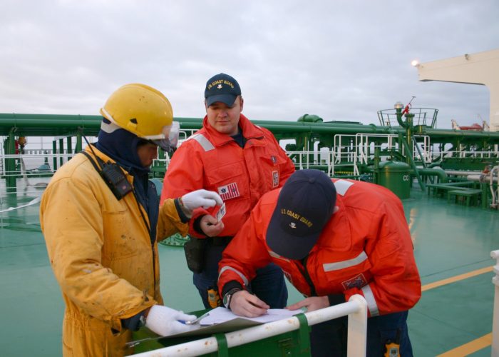 one man wearing yellow jacket, hat and gloves and two other men wearing orange jacket standing on deck while one is writing on paper