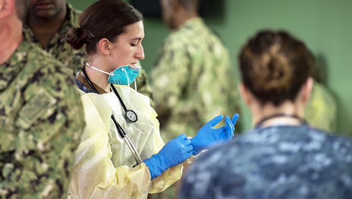 military woman wearing yellow PPE with sleeves, blue gloves, and blue mask with other military members around her
